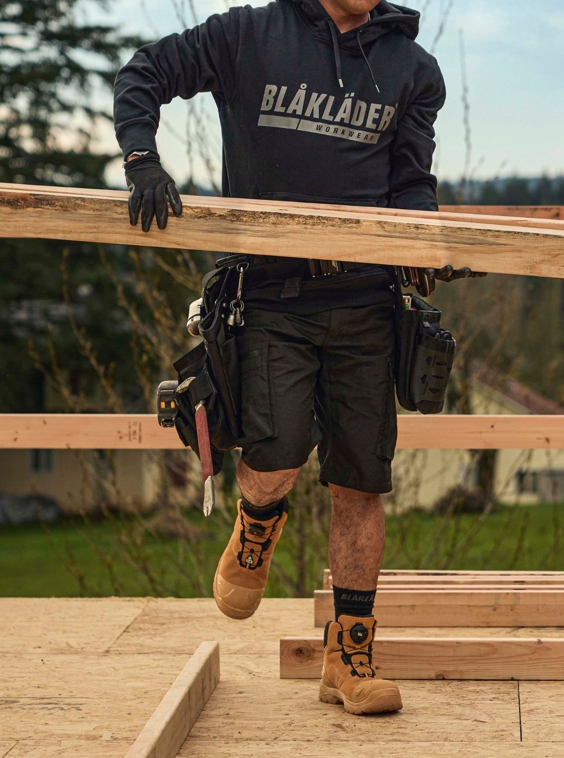 Blaklader Service Shorts with Stretch in Black and Elite Freelock Safety Boots in Wheat worn by carpenter carrying lumber