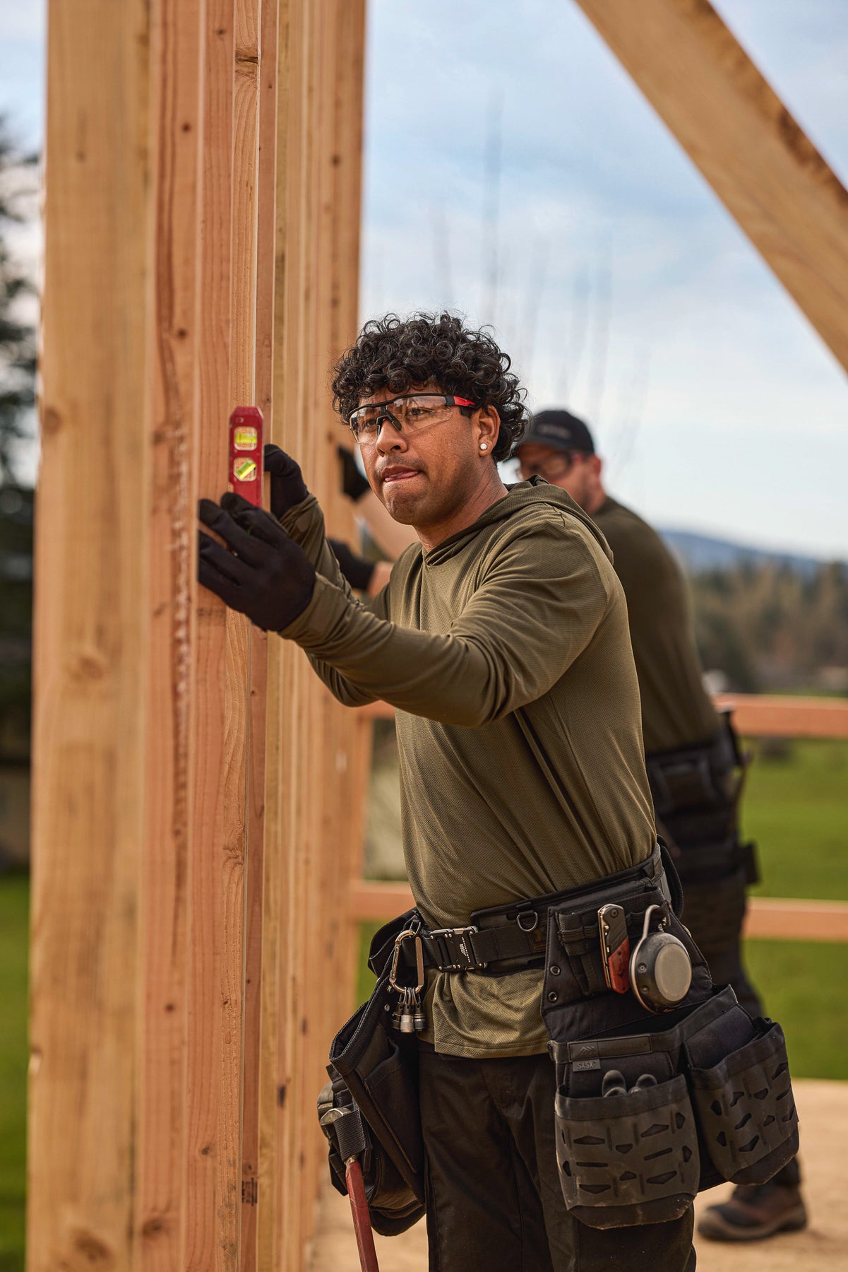 Man in workwear green sun shirt and tool belt using a level on wooden beams outdoors