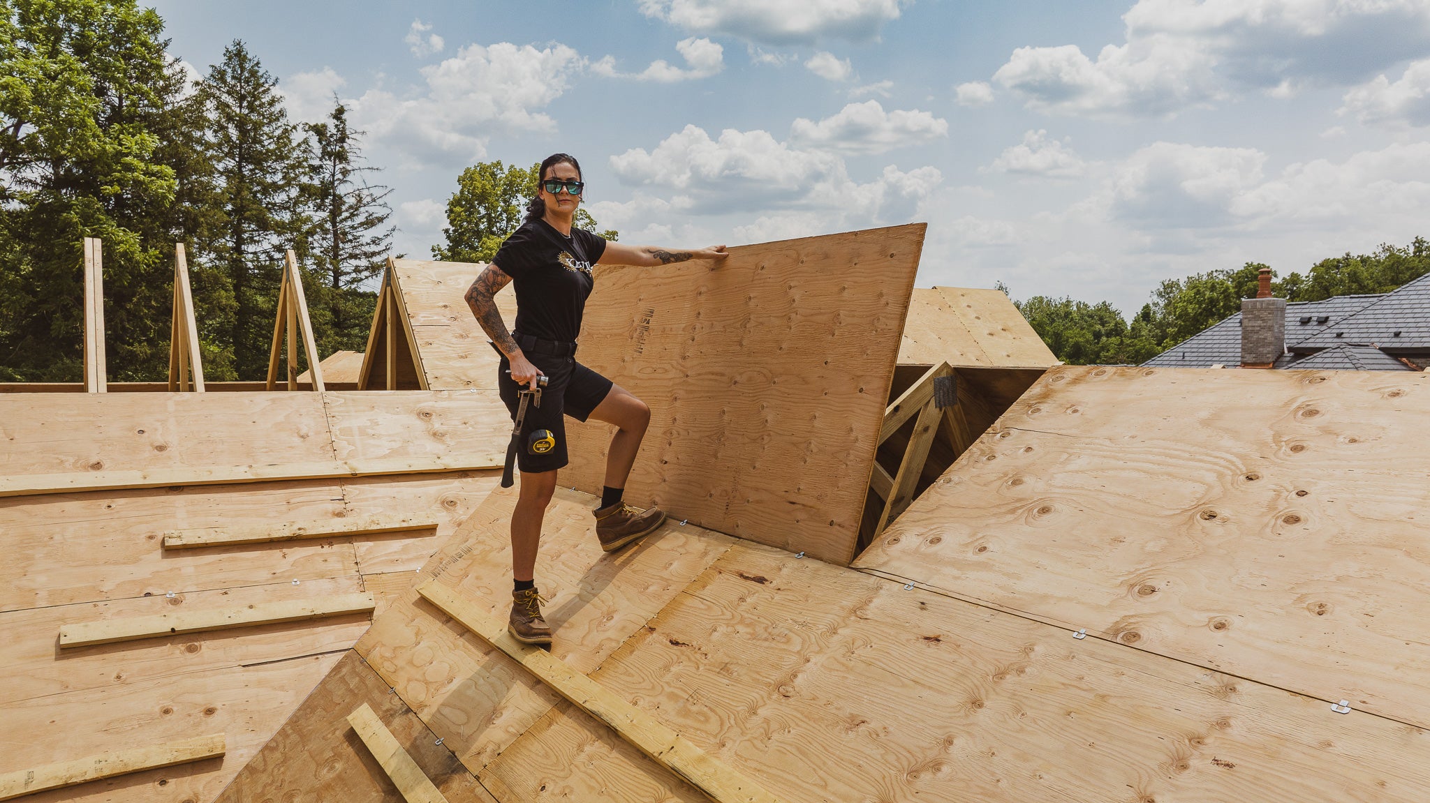 A woman in workwear on a roof holding a wooden panel with construction site and trees in the background